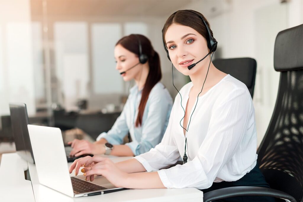 Two women sitting at a desk with laptops and headphones on.